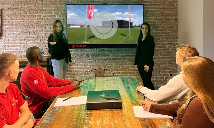 Two young female employees explain the structure of Stadler Holding using a diagram on a large monitor. Four other employees sit at a table and listen attentively.