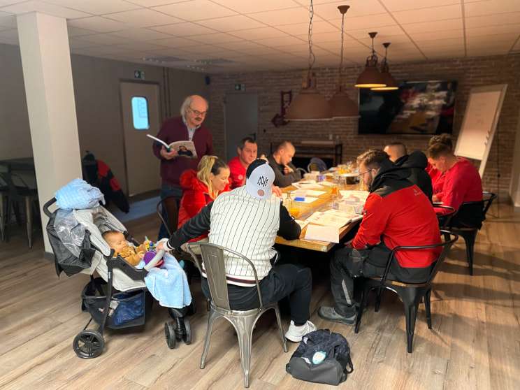 Group of employees at a long table learning German. The teacher stands a bit apart and explains something. A stroller can be seen in the foreground.