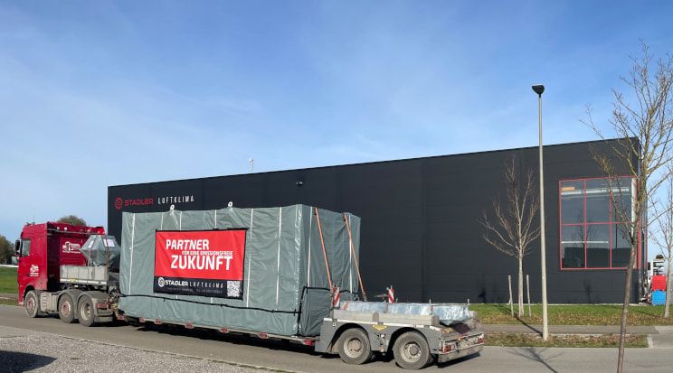 Ein LKW transportiert ein verpacktes Lüftungsmodul ab. Auf der Verpackung ist ein Banner mit der Aufschrift "Partner für eine Emissionsfreie Zukunft" zu sehen, das die Stadler Luftklima bewirbt.