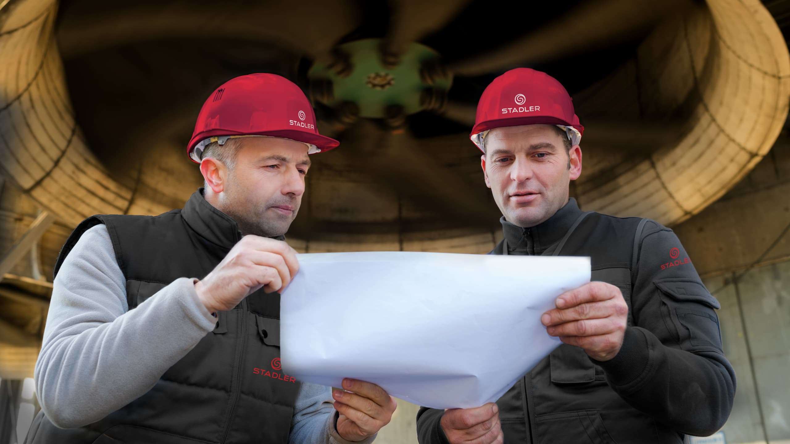 Two men holding a blueprint under a ventilation turbine