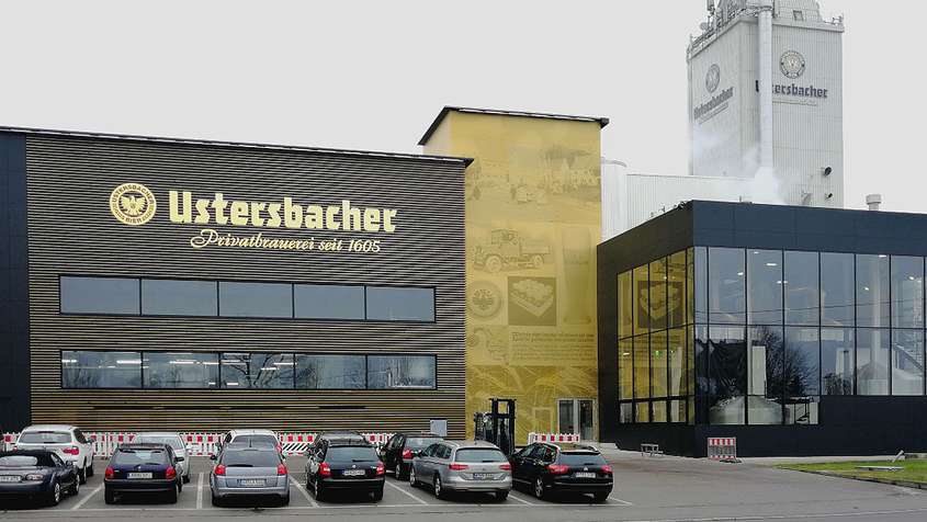 Gold-braune Gebäude hinter einem Parkplatz. Eine Fassade an der rechten Seite trägt die Aufschrift „Ustersbacher“
