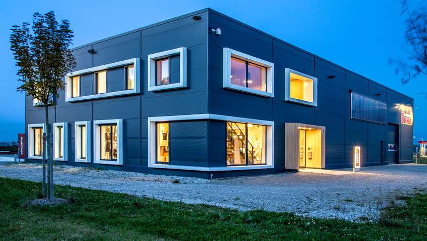 A rectangular building constructed from dark grey panels with lit up windows and wide, white window frames