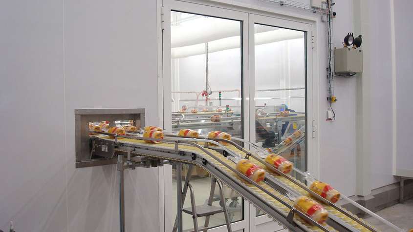 A conveyor belt transporting packaged bread through a white wall with a large window looking into a room with more conveyor belts