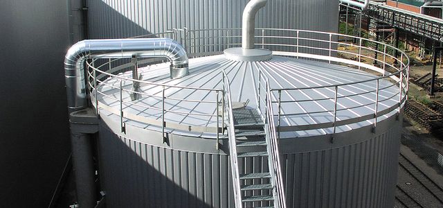 A large silo with a metal staircase. Some steam is flowing out of a pipe at the top of the silo