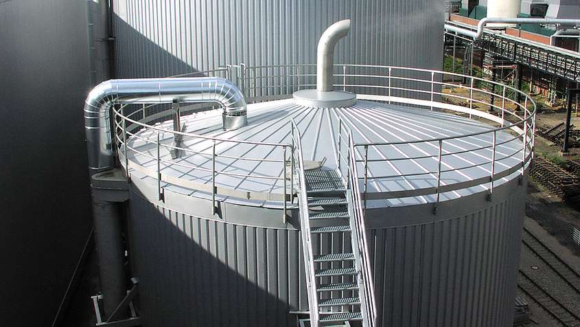 A large silo with a metal staircase. Some steam is flowing out of a pipe at the top of the silo