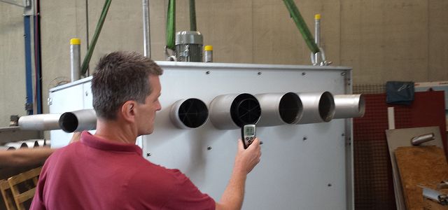 Man in a red shirt measuring something standing in front of a metal box with lots of air outlets