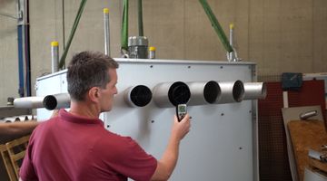 Man in a red shirt measuring something standing in front of a metal box with lots of air outlets