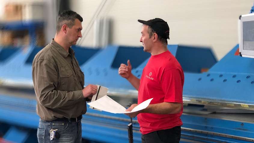 Two men holding documents and talking in front of a machine