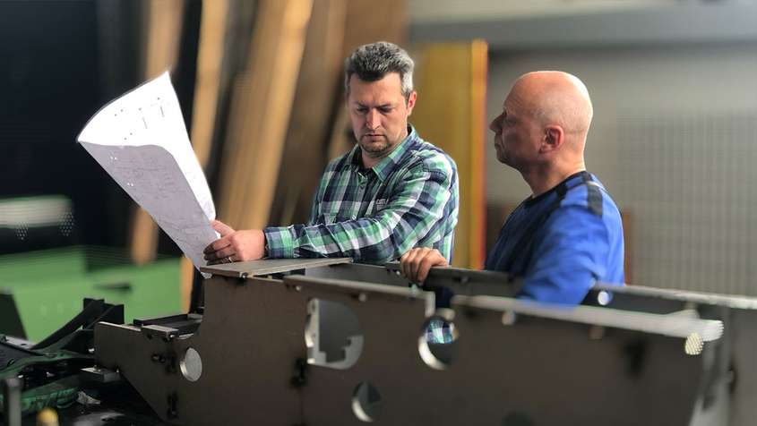 Two men looking at a blueprint. They’re standing behind a large metal workpiece