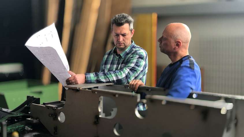 Two men looking at a blueprint. They’re standing behind a large metal workpiece