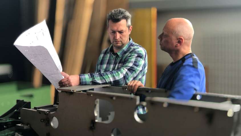 Two men looking at a blueprint. They’re standing behind a large metal workpiece