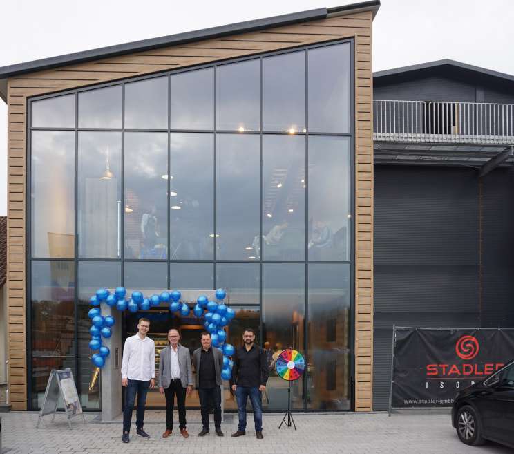 Eine Gruppe Menschen steht vor einem modernen Gebäude, dessen Eingang mit blauen Luftballons verziert ist.