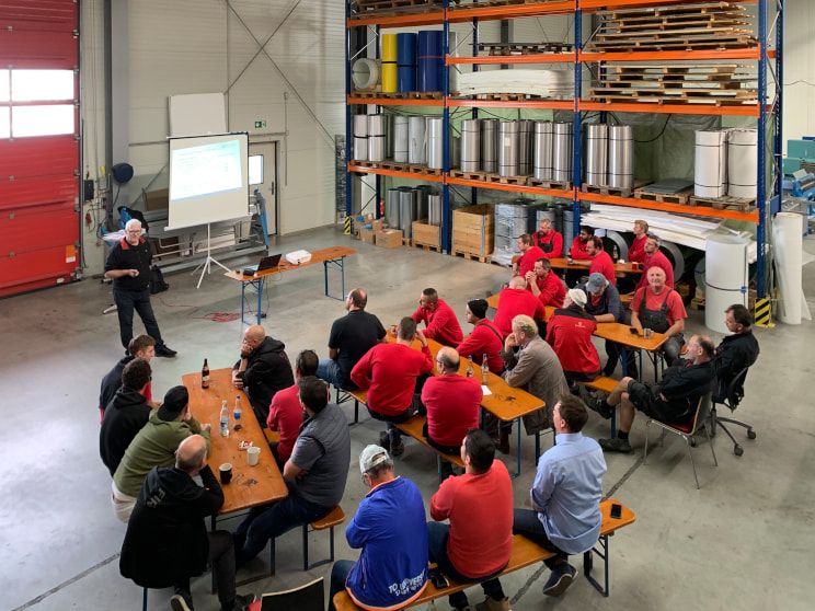 A crowd sits on beer benches in a warehouse listening to a speaker present something on a portable screen.